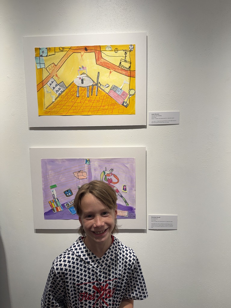 A child with short hair smiles, standing next to two framed paintings on a white wall.