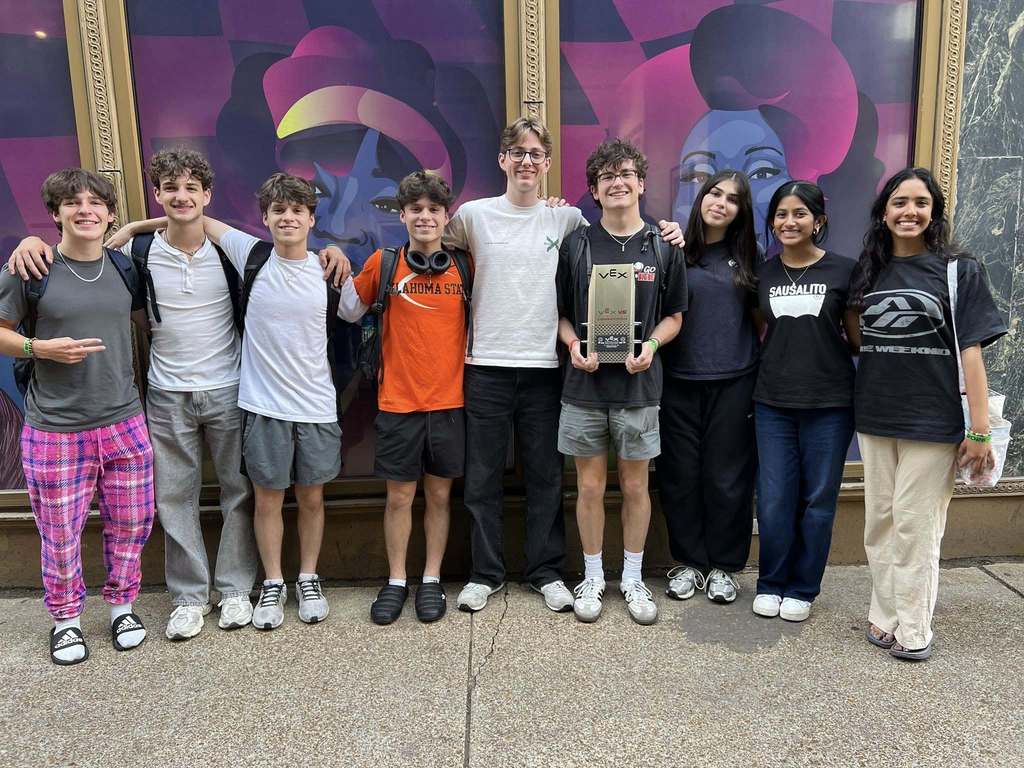 Nine young individuals stand in front of a mural, smiling and posing for a photo. They wear casual clothing, including sneakers.