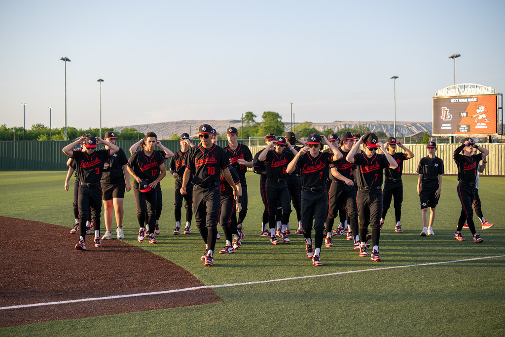 A baseball team walks on a field, wearing black uniforms. A large scoreboard is in the background.