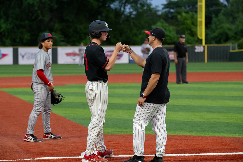 Three individuals on a baseball field. Two in black jerseys, one in gray, shake hands. Behind them, a field with a banner and umpire.
