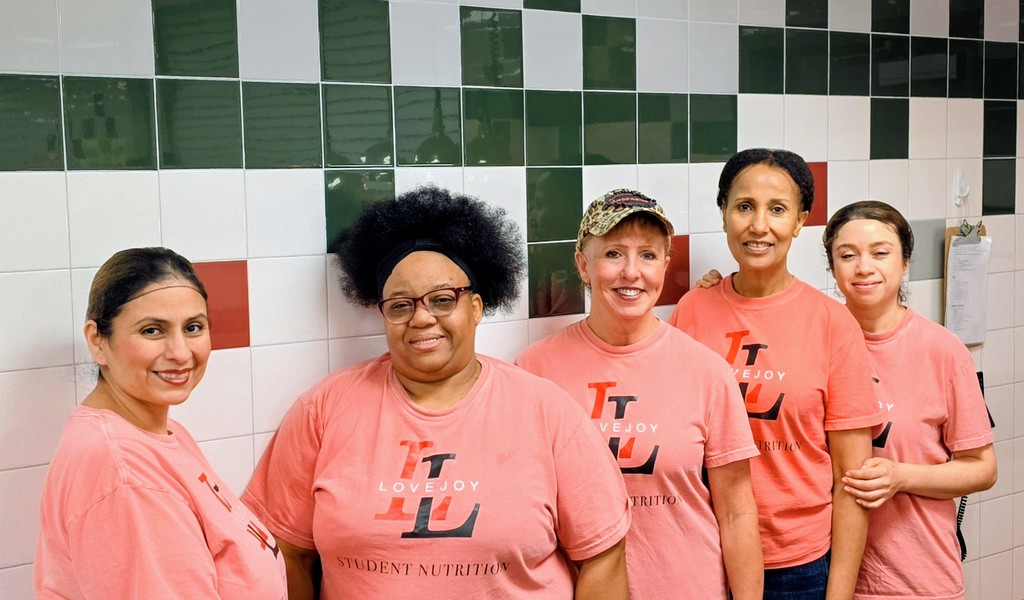 Five women in matching pink shirts stand in a kitchen. They are smiling and appear to be posing for a photo.
