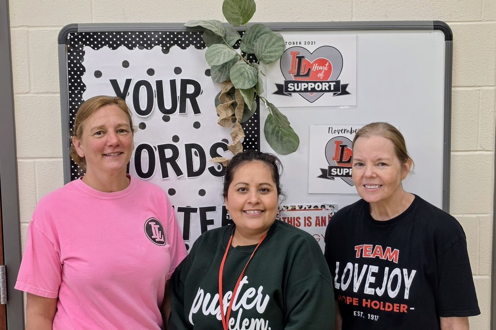 Three women in T-shirts stand in front of a bulletin board with green leaves and motivational text.
