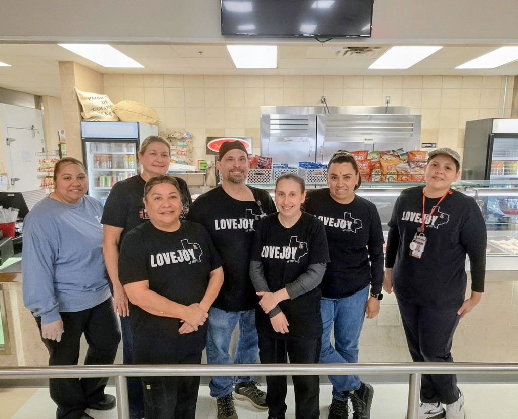 A group of people in black shirts are standing together in a kitchen. A metal barrier is in front.