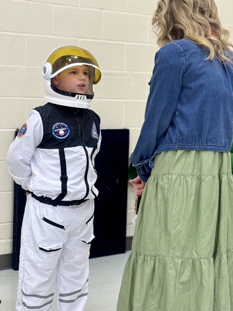 A child in a space suit and helmet talks to an adult in a blue jacket and green skirt.