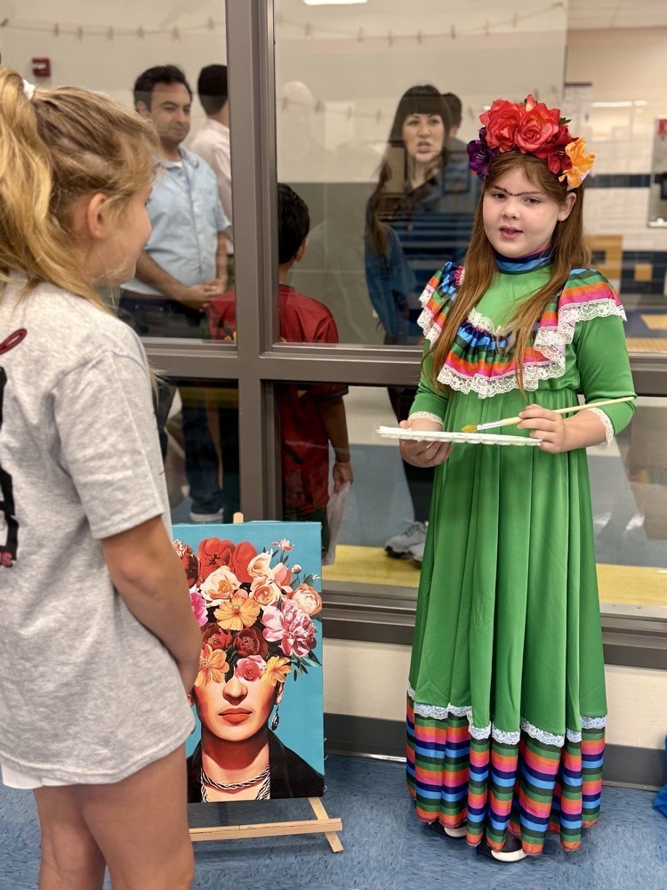 Two girls inside a room. One girl holds a paint palette and the other looks at a painting.