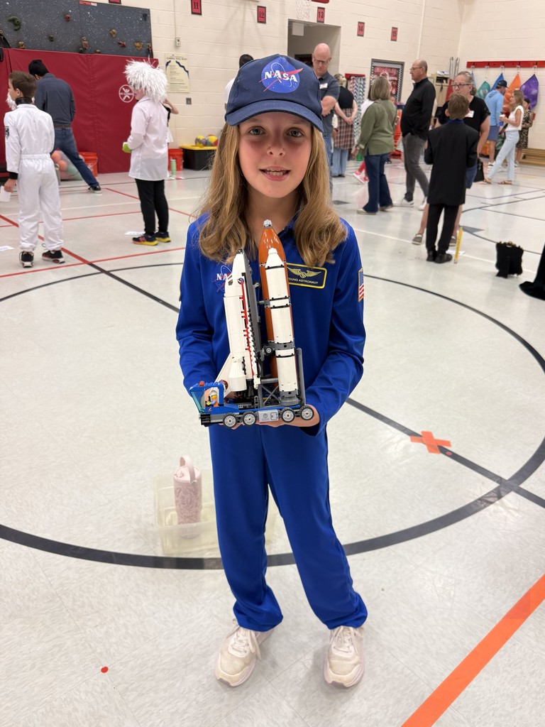 A child in a blue costume holds a space shuttle model at an event. Behind her, people walk on a gym floor.