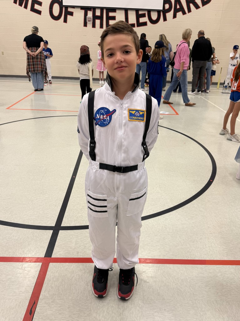 A child in a NASA space suit stands in a gymnasium, with others dressed in various costumes around.