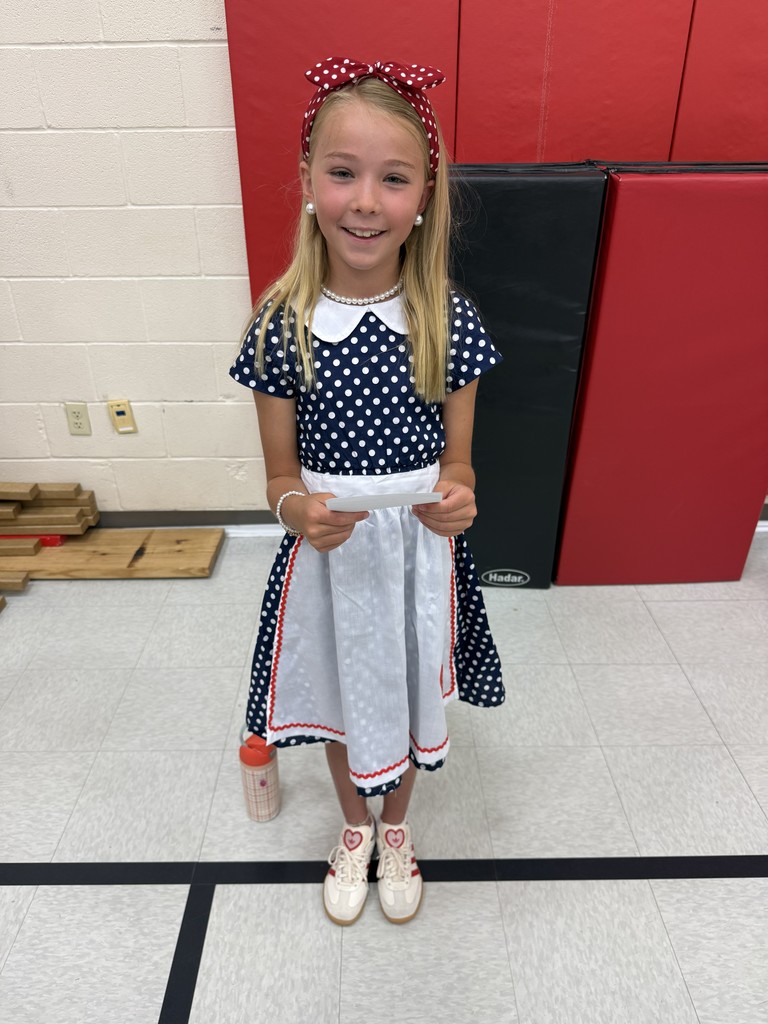 A young girl in a polka dot dress with a red bow stands in a school hallway.