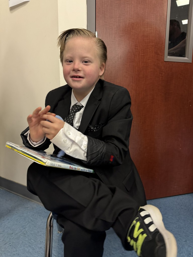 A child in a suit sits on a stool and holds a book, smiling at the camera.