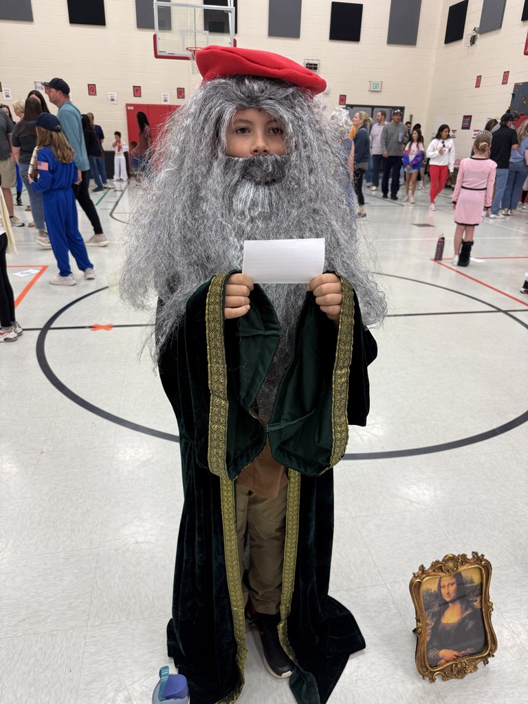 A child dressed as Gandalf holds a paper, standing in a gymnasium with people walking around.