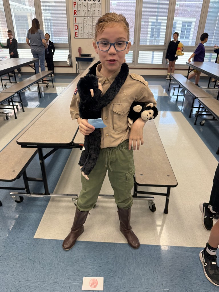 Child in scout uniform, glasses, and boots. Carrying a stuffed animal and holding a card. Background includes tables and students.