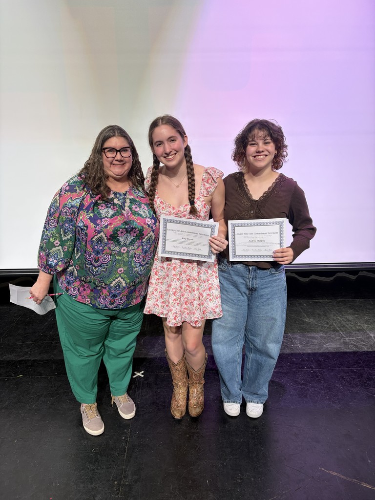 Three women stand on a stage with certificates; one holds a paper, another has braids, and the last wears boots. A screen behind them shows a purple hue.