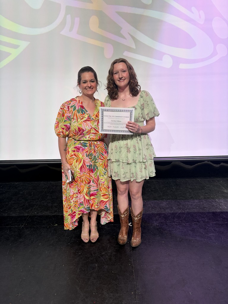Two women on stage, one in a floral dress and the other in a green dress, holding a certificate.