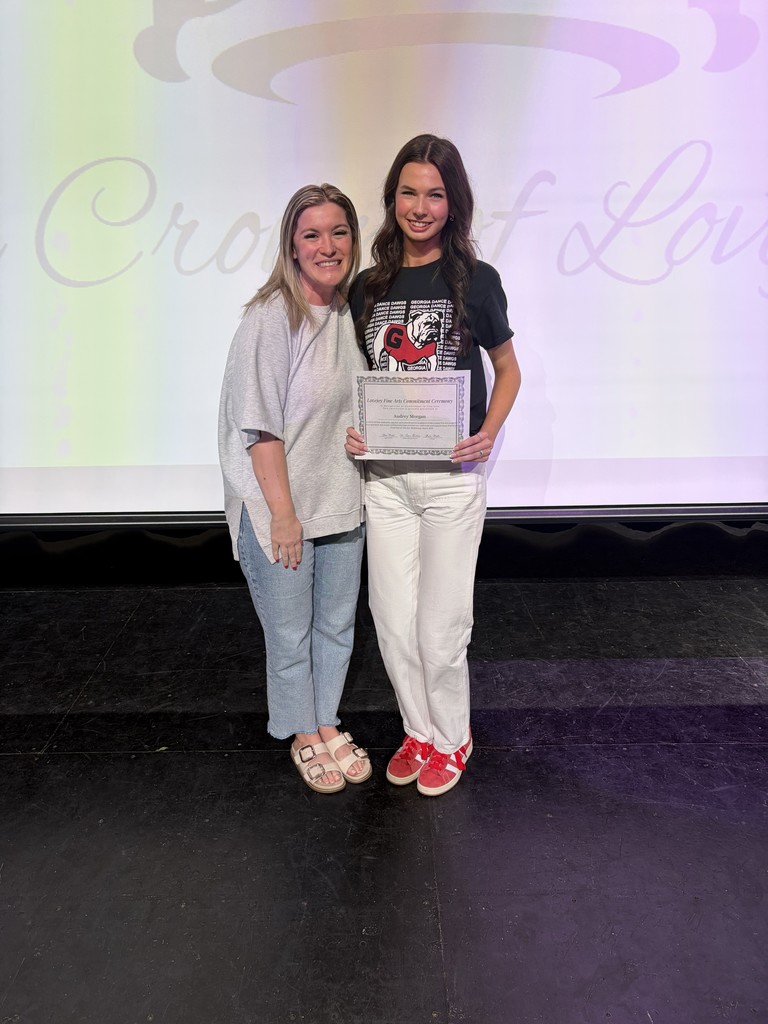 Two women stand on a stage with a backdrop displaying "Crown of Love". One holds an award, wearing a black t-shirt and red sneakers. The other wears a gray top, blue jeans, and sandals.