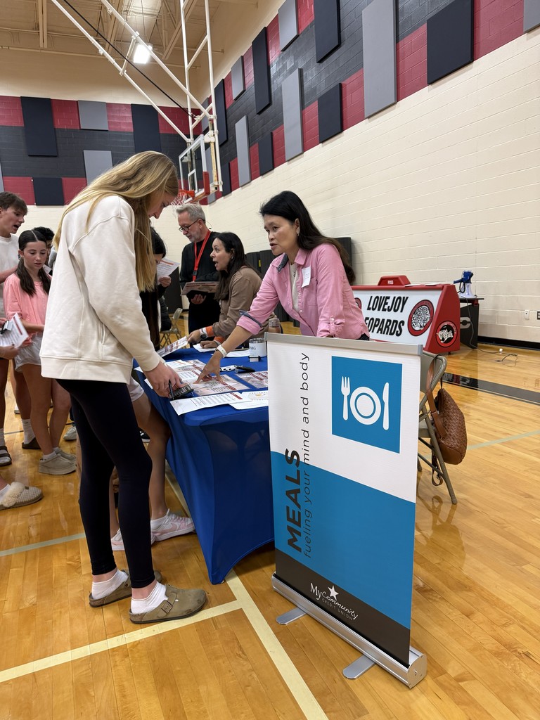 A woman stands at a table with a blue tablecloth, reading materials. Another woman stands behind her. A sign reads "Meals on Wheels."