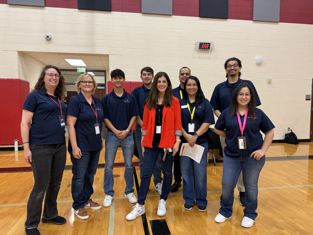 Eleven people stand in a school gym, smiling and posing for a photo, with a basketball court visible.