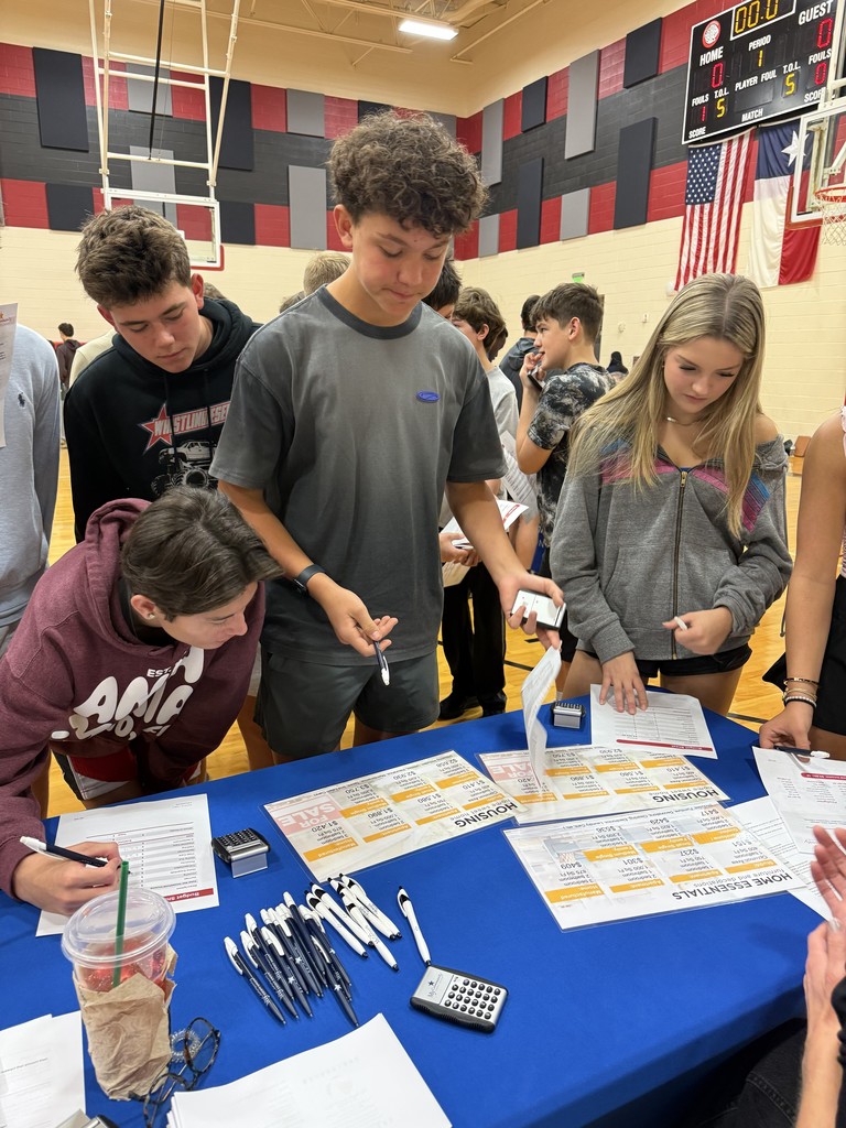 Several young people gather around a table in a gymnasium with papers, pens, and a calculator.