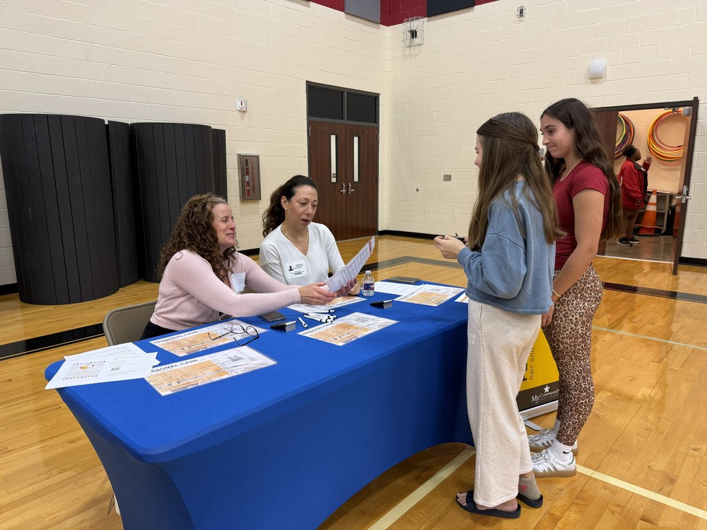 Four women at a table in a gymnasium. Papers are on the table. One woman is seated and talking to two standing women.