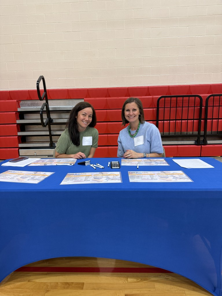 Two women sitting at a blue table, smiling. Papers, pens, and a phone are on the table. Behind them, a red bleacher.