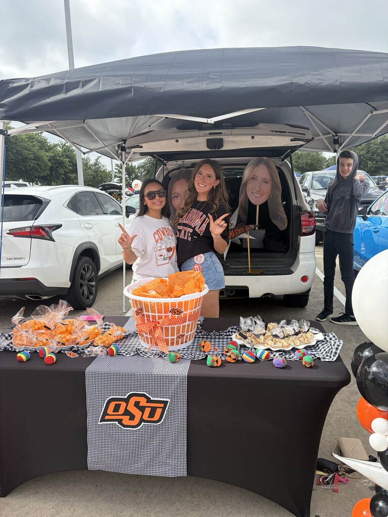 Three people stand behind a table with food in an outdoor tent. The table has balloons and a banner.