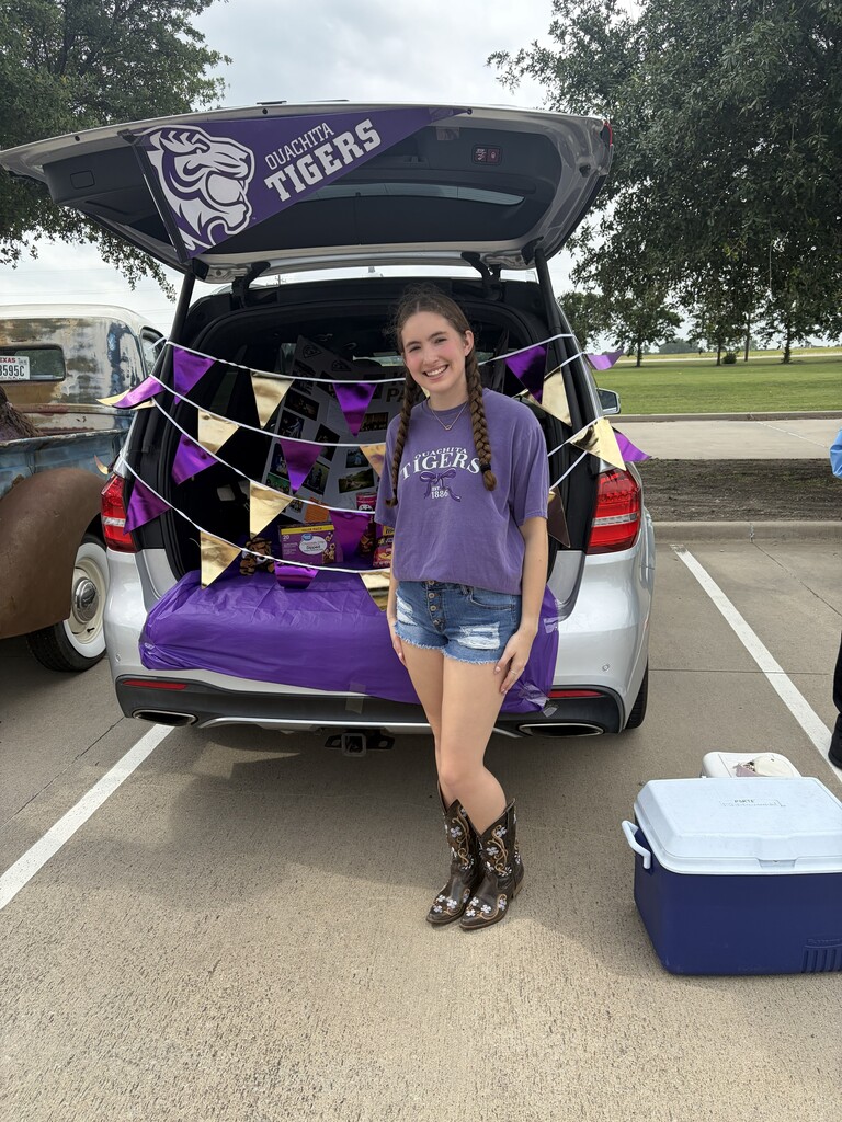 A girl wearing a purple shirt and denim shorts stands next to a car trunk decorated with a team banner.
