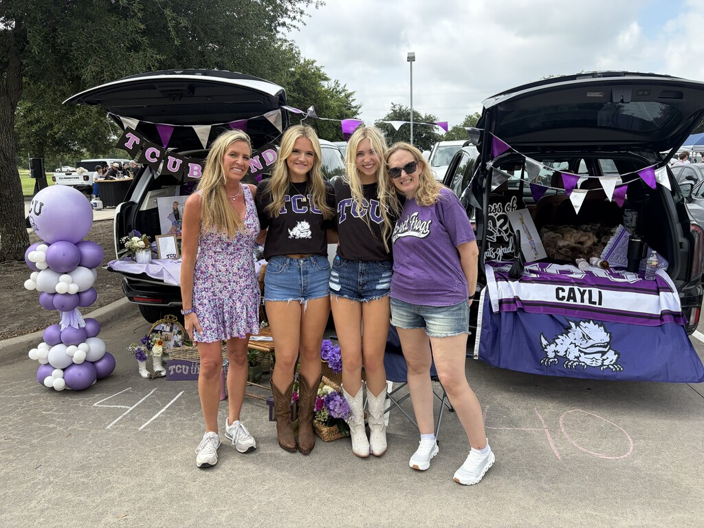 Four women in matching outfits stand by their cars in a parking lot. Purple balloons and table decorations are in the background.