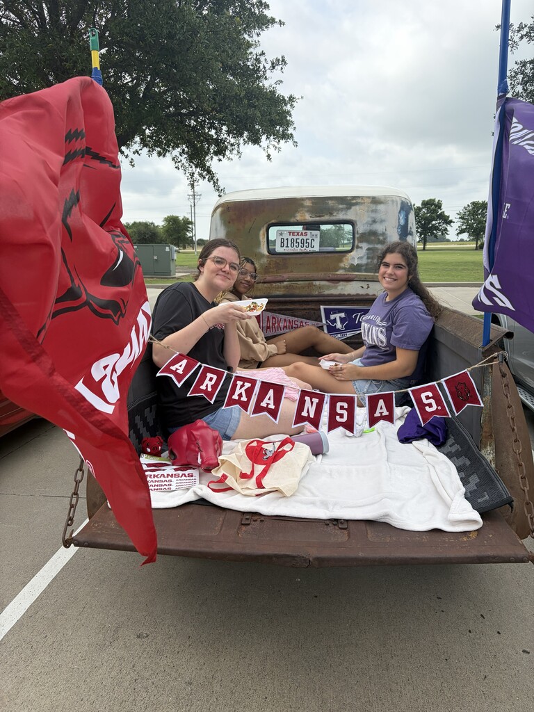 Three people sit in a pickup truck bed with a banner reading "Arkansas" and holding food and drinks.