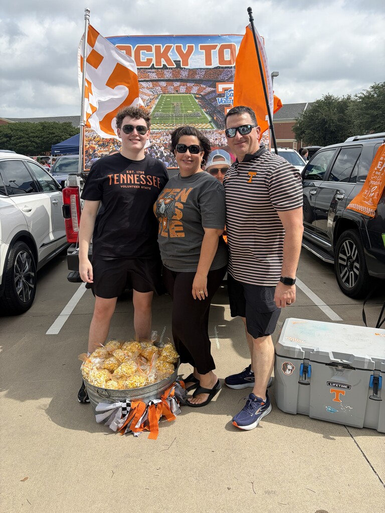 Three people stand next to a bucket of popcorn in a parking lot. A car is parked behind them, and a cooler is nearby.