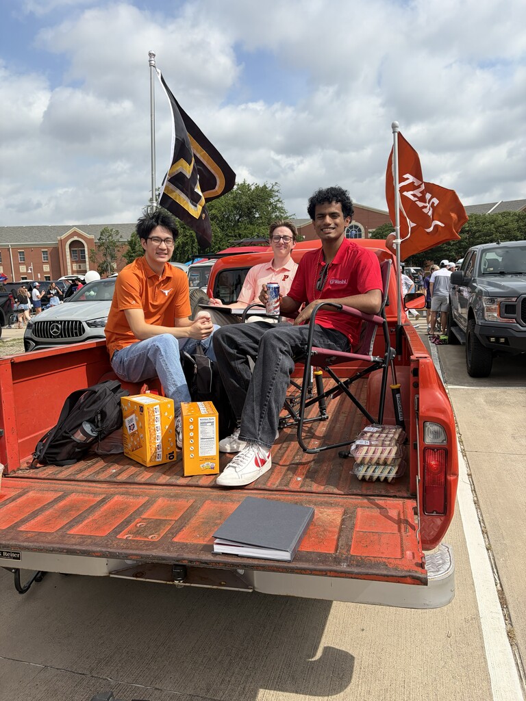 Three people sit in a red truck bed. Two are seated, one standing. Flags and books are present.