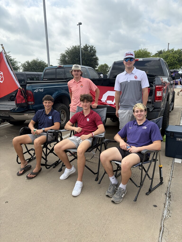 Five young men wearing hats and sneakers sit on chairs in a parking lot. Vehicles are parked behind them.