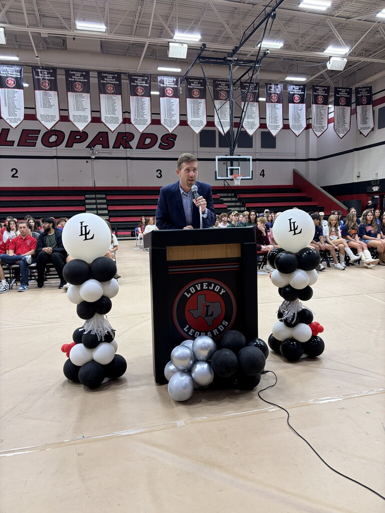 Person speaking at a podium in a gymnasium, surrounded by balloons and banners. A crowd of people sits in the background.