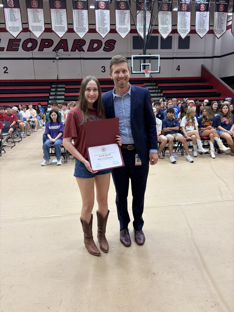 Woman holding certificate stands next to man in suit, indoors. Behind them, a crowd sits in bleachers. Banner reads "LEOPARDS" on wall.