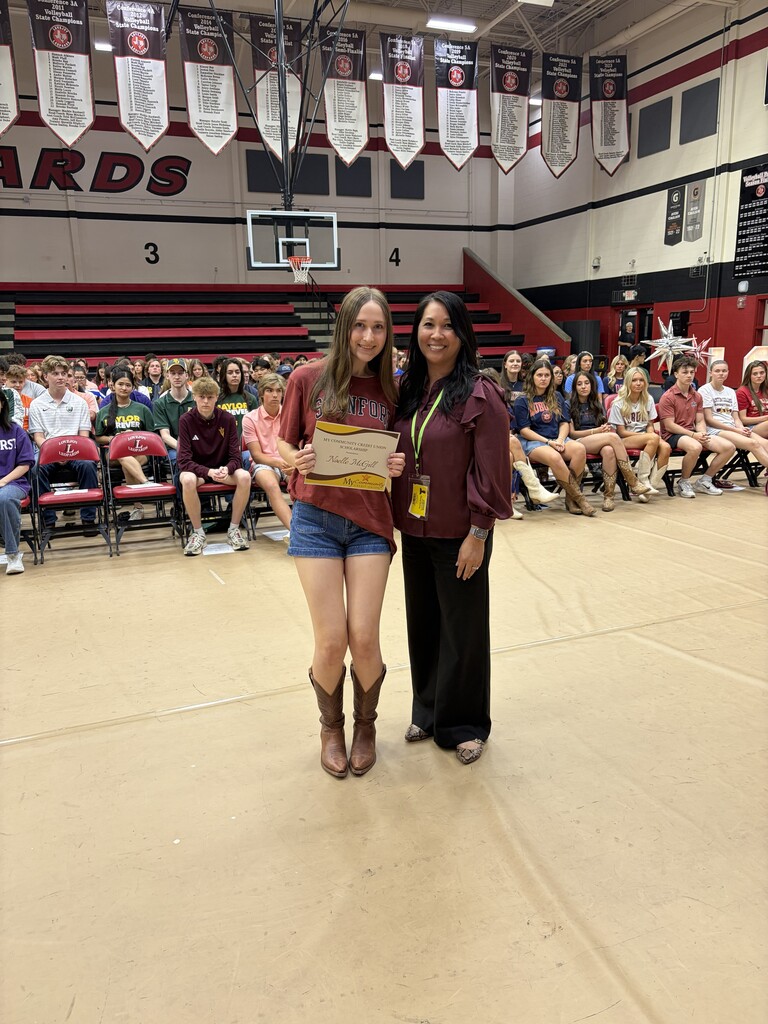 Two women stand in a school gym, one holding a certificate. Behind them, students sit on bleachers.