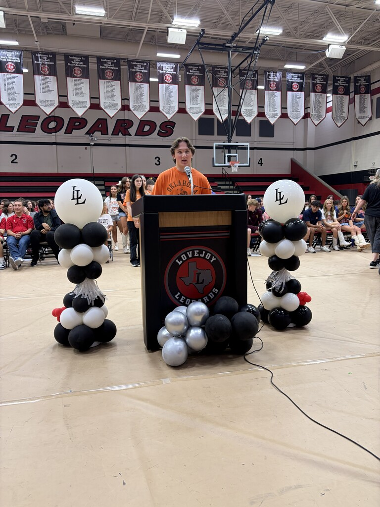 A person speaks from a podium in a gymnasium, surrounded by balloons. Banners are displayed above.