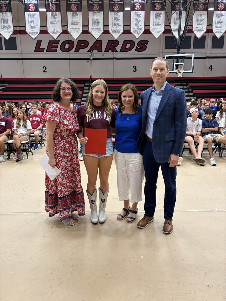 Five people stand together in front of a wall with "Leopards" written on it. One holds a red folder.