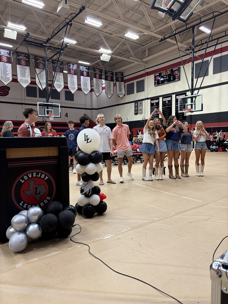 A gymnasium with a basketball court, a podium, and a large balloon display. People are gathered, some are taking pictures.