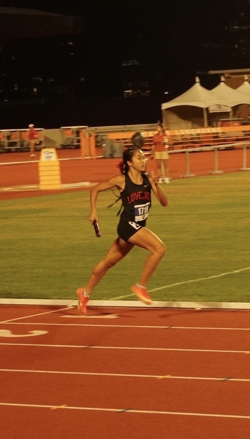 A woman in a black tank top runs on a track, holding a baton. Behind her, a grassy field and tents.