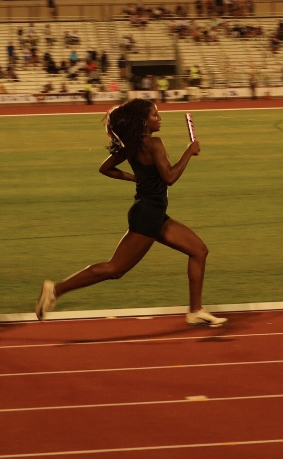 A woman runs on a track at night. She holds a baton. In the background, spectators are seated.