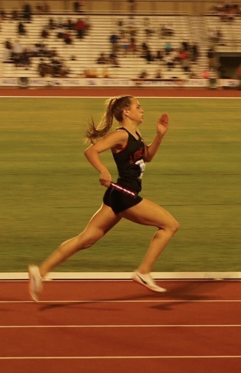 A woman in a black singlet runs on a track, holding a baton. Behind her, a green field and spectators.