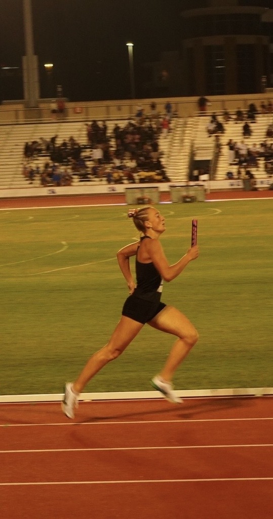 A female runner on a track with a baton in her right hand, running on a track with spectators in stands.