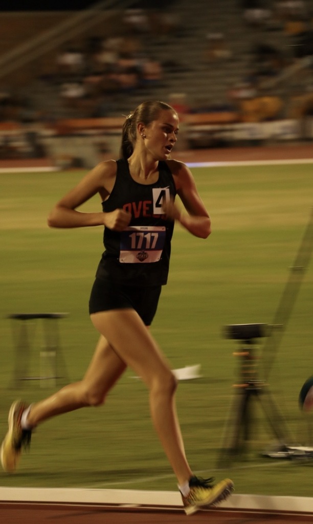 A female athlete runs on a track, wearing a black tank top with "OVE" and number 4.