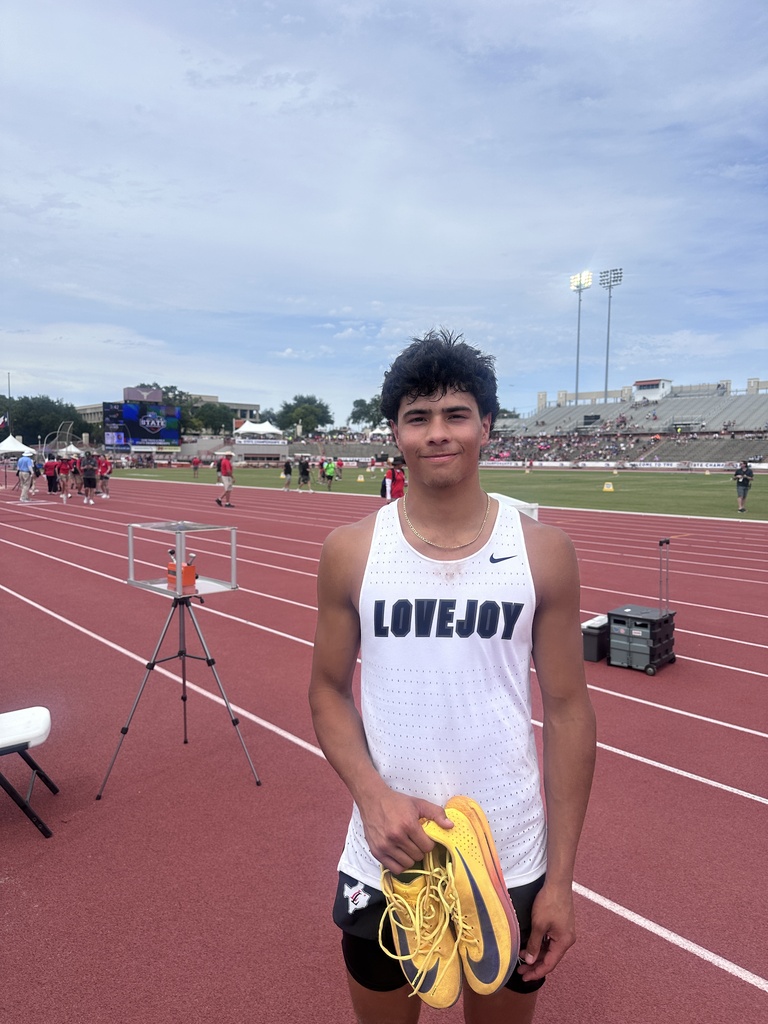 Man in white tank top reading LOVEJOY holds yellow shoes on a red track with a stadium in the background.