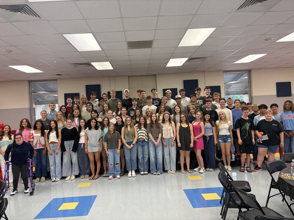 A large group of people in casual clothing standing in rows inside a room with white walls and ceiling lights.