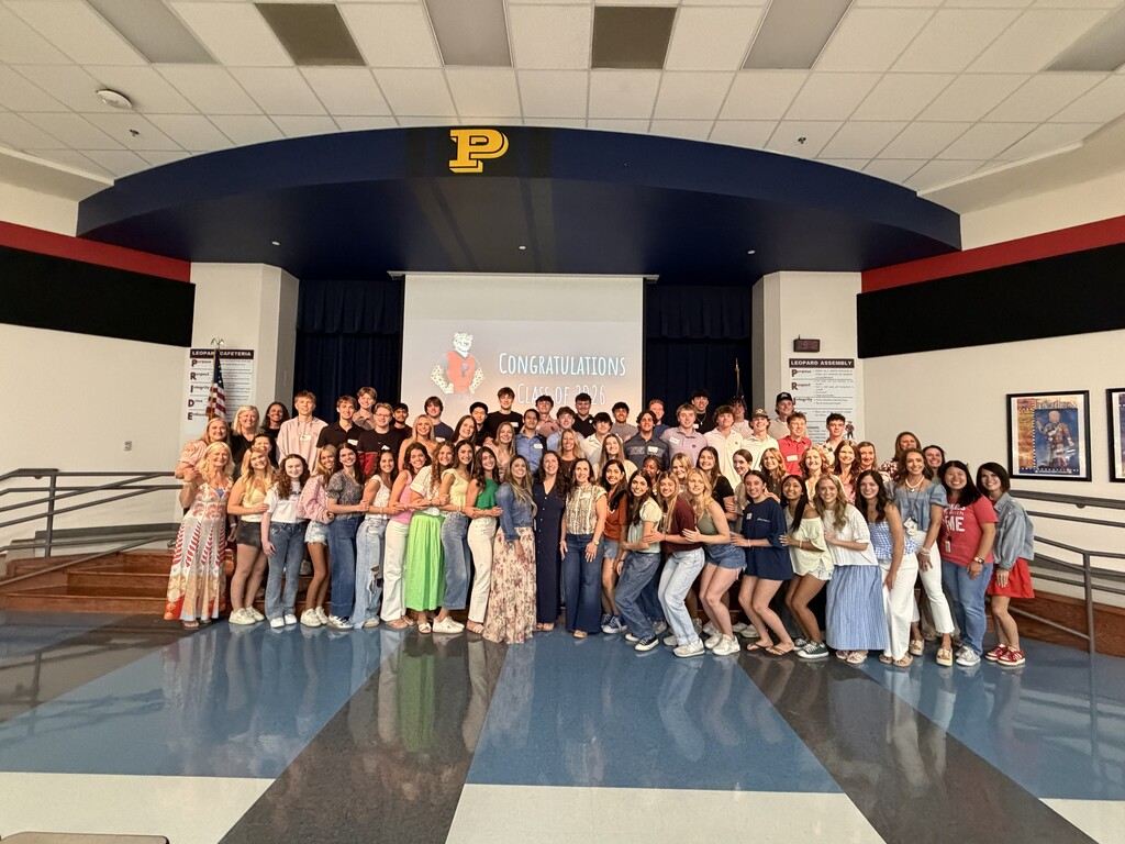 A group of people standing in a room with a stage, projector screen, and "P" symbol.