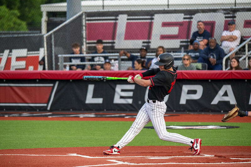 Baseball player in black and white uniform swings green bat on field with red dirt; crowd in stands behind wall.