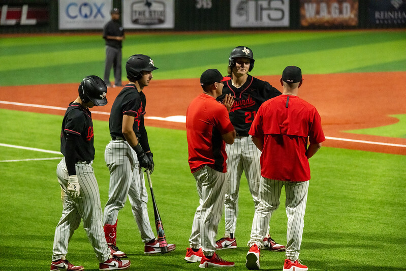 Players in black jerseys huddle on a baseball field. Some hold bats. Two others wear red shirts.