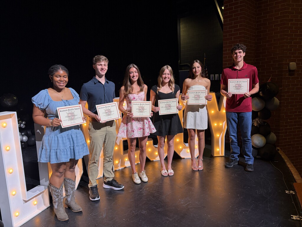 Six students stand on stage, holding certificates, near large lighted letters spelling "LOVE."
