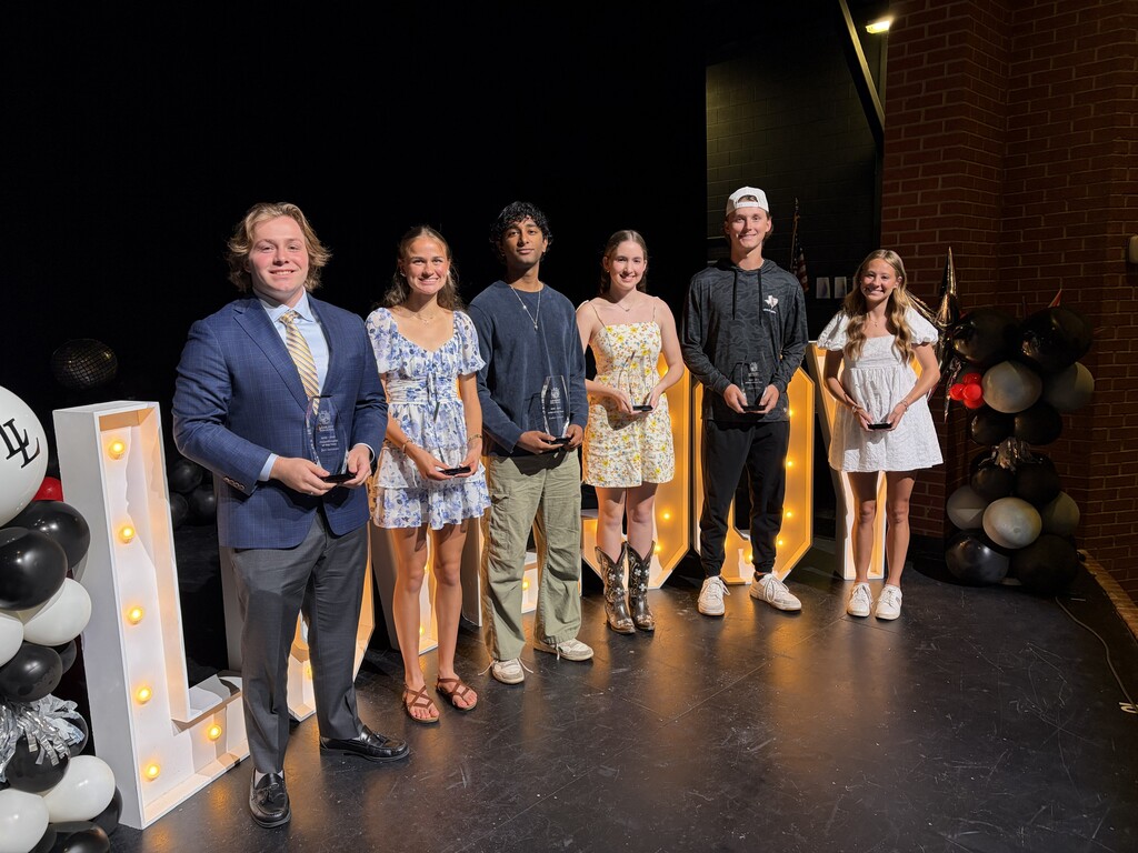 Six individuals in formal attire stand on stage, holding awards, with balloons and lights forming the letter "L".