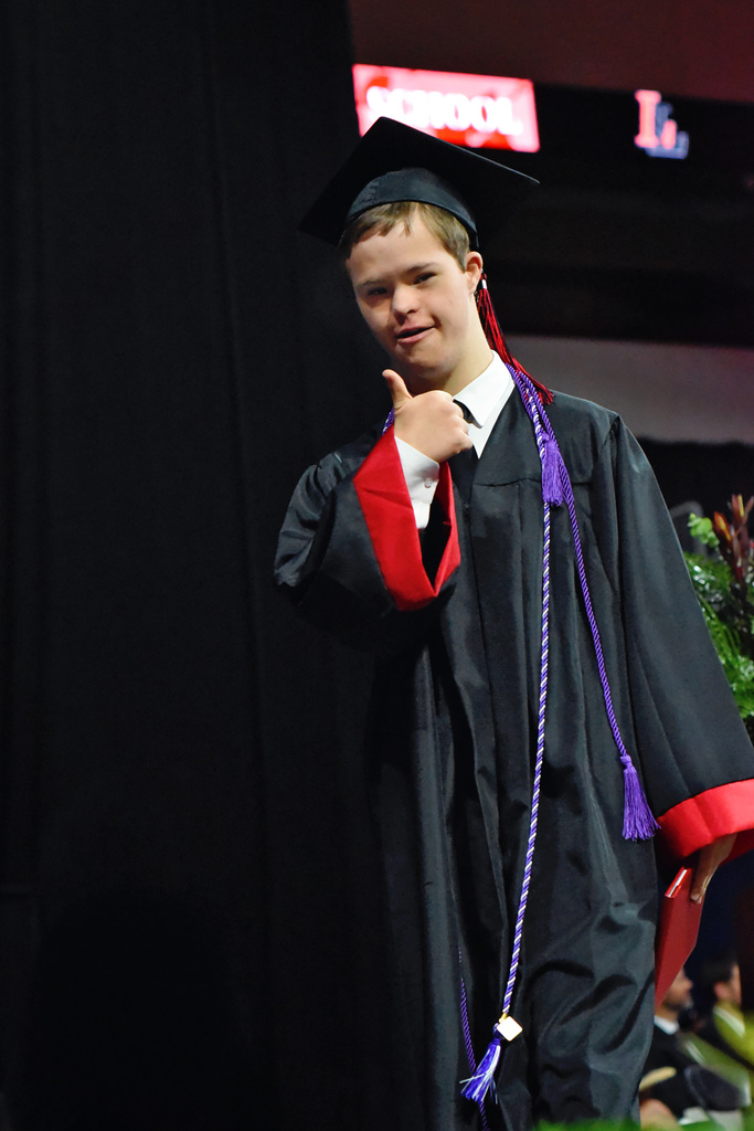 A young man in a graduation gown and cap, standing on stage, gives a thumbs-up gesture.