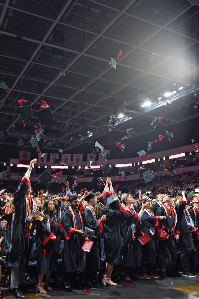 Graduates in black gowns and caps throw them in the air at a ceremony in an indoor stadium.